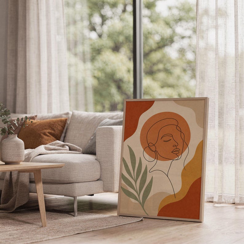 Abstract line art face print leaning on the floor beside a cream armchair next to large garden windows.