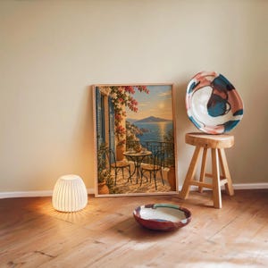 Mediterranean balcony wall art print leaning against a warm beige wall on a wooden floor beside a stool and a glowing lamp.