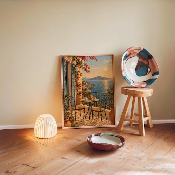 Mediterranean balcony wall art print leaning against a warm beige wall on a wooden floor beside a stool and a glowing lamp.