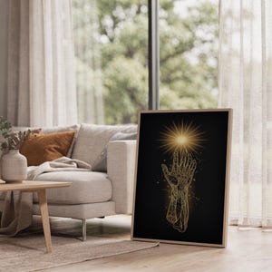 Celestial hand wall art print leaning on the floor beside a cream armchair next to large garden windows.