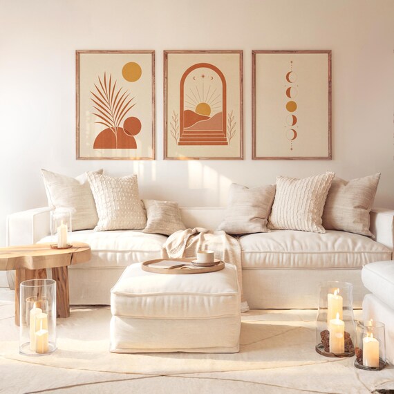 Boho wall art set of 3 with warm terracotta tones and celestial sun prints hung above a large beige sectional couch with pillows and candles in a bright, airy minimalist living room environment.