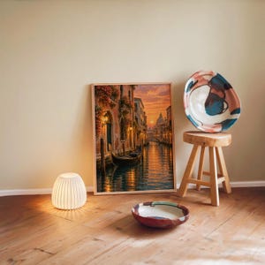 Venice canal sunset wall art print leaning against a warm beige wall on a wooden floor beside a stool and a glowing lamp.