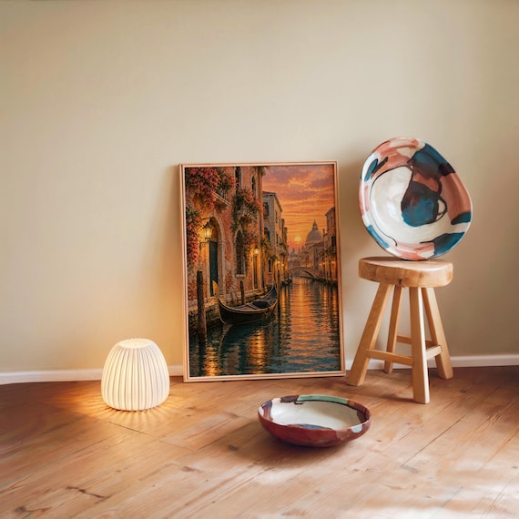 Venice canal sunset wall art print leaning against a warm beige wall on a wooden floor beside a stool and a glowing lamp.