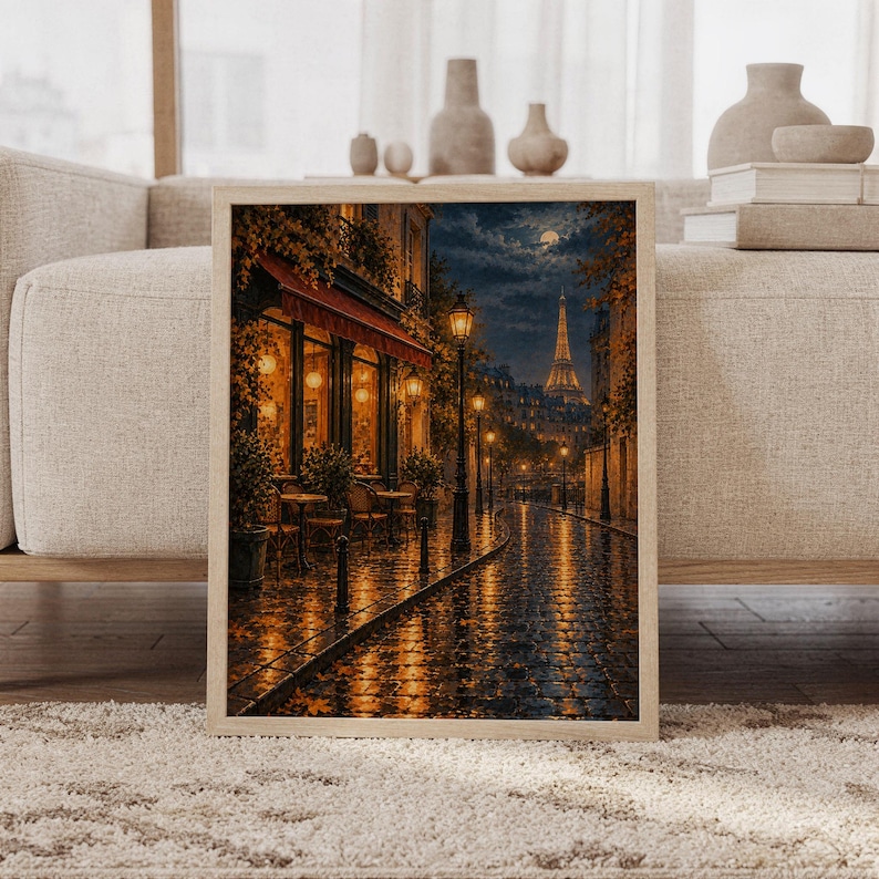 Paris eiffel tower wall art print featuring deep midnight blue skies, warm amber streetlights, a glowing Eiffel Tower, rain-slick cobblestones, and an inviting Parisian cafe in a painterly cityscape style, framed and leaning against a beige sofa on a textured rug in a bright Scandinavian living room.