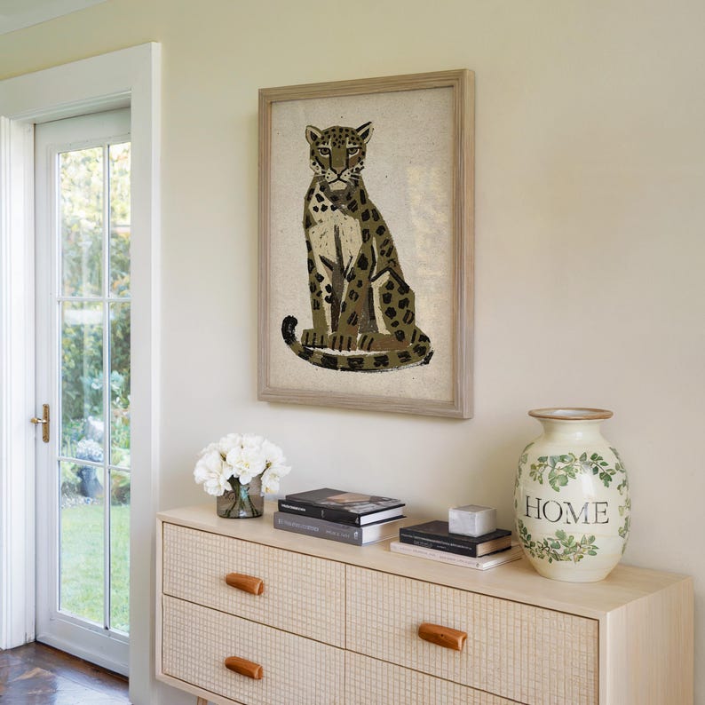 A framed print of a sitting leopard with dark green and brown spots on a light background, placed on a light wood dresser with books and a vase that says "HOME."