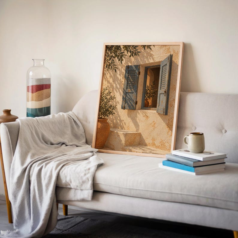 Mediterranean blue shutter wall art print leaning on a cream sofa against a beige wall beside books and a mug.
