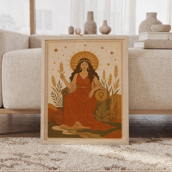 Celestial goddess wall art print featuring warm terracotta, ochre, olive green, and cream tones, a serene goddess seated beneath a radiant sun with stars, wheat, and a feminine Venus symbol, framed and leaning against a beige sofa on a textured rug in a bright Scandinavian living room.