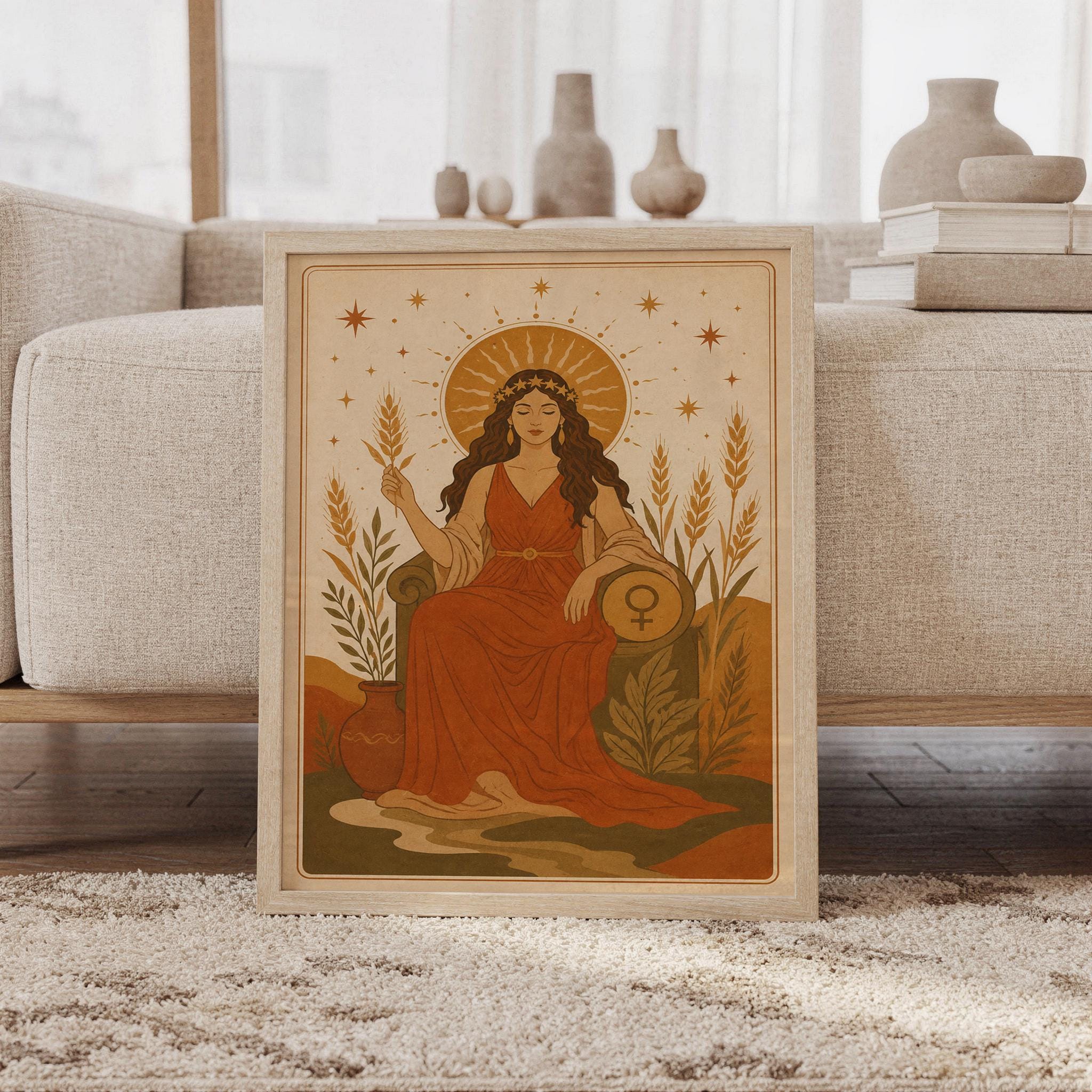 Celestial goddess wall art print featuring warm terracotta, ochre, olive green, and cream tones, a serene goddess seated beneath a radiant sun with stars, wheat, and a feminine Venus symbol, framed and leaning against a beige sofa on a textured rug in a bright Scandinavian living room.