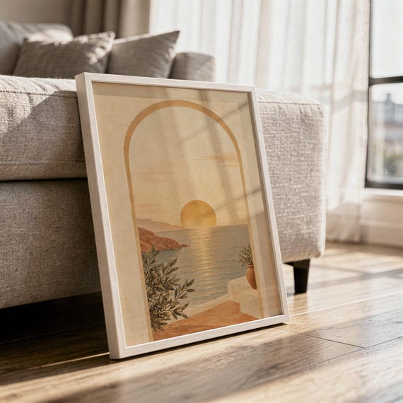 Mediterranean sunset arch print in a white frame leaning against a gray sofa on a sunlit wooden floor.