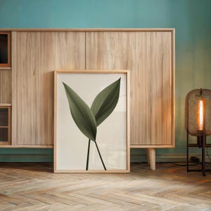 A framed print of two dark green leaves on a light background rests on a wooden floor in front of a light wood cabinet and a metal lamp.
