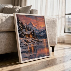 Winter cabin mountain wall art print in a white frame leaning against a gray sofa on a sunlit wooden floor.