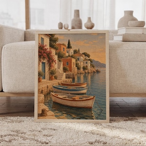 Mediterranean coastal village wall art print featuring warm terracotta homes, blue shutters, flowering bougainvillea, cypress trees, and wooden boats floating on calm blue water, framed and leaning against a beige sofa on a textured rug in a bright Scandinavian living room.