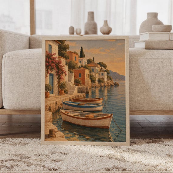 Mediterranean coastal village wall art print featuring warm terracotta homes, blue shutters, flowering bougainvillea, cypress trees, and wooden boats floating on calm blue water, framed and leaning against a beige sofa on a textured rug in a bright Scandinavian living room.