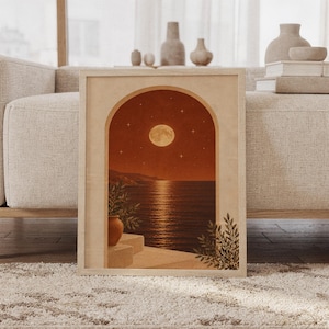 Moonlit Mediterranean arch wall art print of a glowing full moon and stars over a calm sea and coastal cliffs, framed by an arch with potted plants in terracotta, cream and olive tones, framed and leaning against a beige sofa on a textured rug in a bright Scandinavian living room.