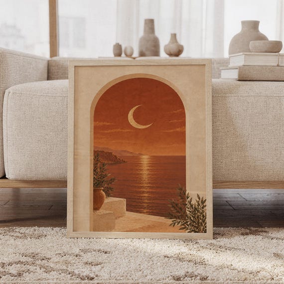 Mediterranean moon arch wall art print of a crescent moon above a calm coastal sea framed by a sun-washed arch in terracotta, burnt sienna, sand and olive tones, framed and leaning against a beige sofa on a textured rug in a bright Scandinavian living room.