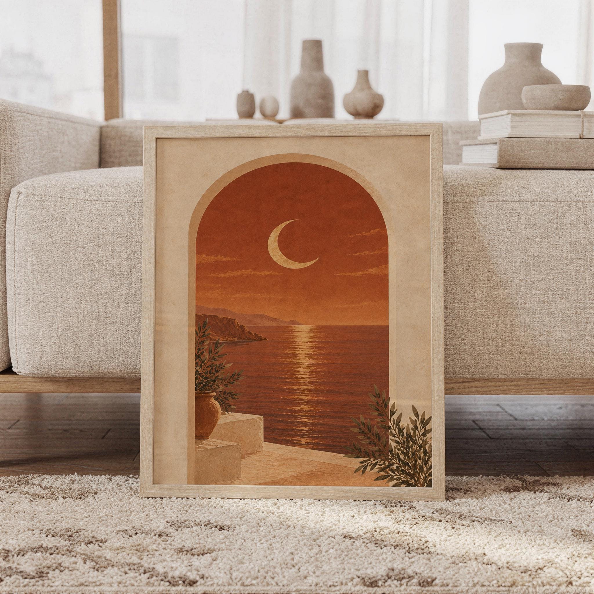 Mediterranean moon arch wall art print of a crescent moon above a calm coastal sea framed by a sun-washed arch in terracotta, burnt sienna, sand and olive tones, framed and leaning against a beige sofa on a textured rug in a bright Scandinavian living room.