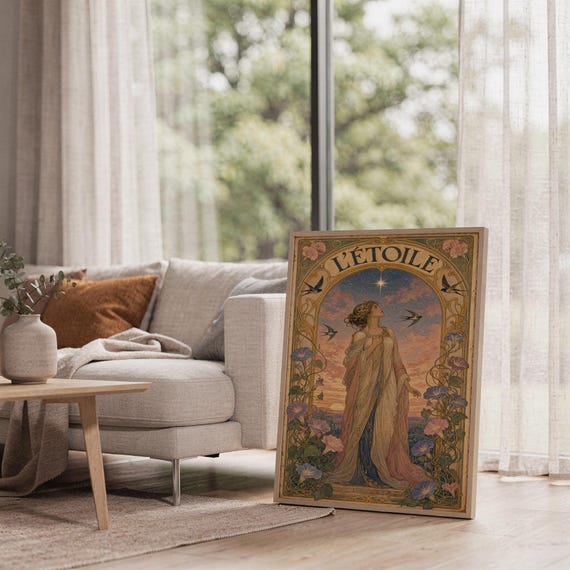An art nouveau L Etoile print in a light wood frame leaning on a wooden floor in a sunlit room with a sofa.