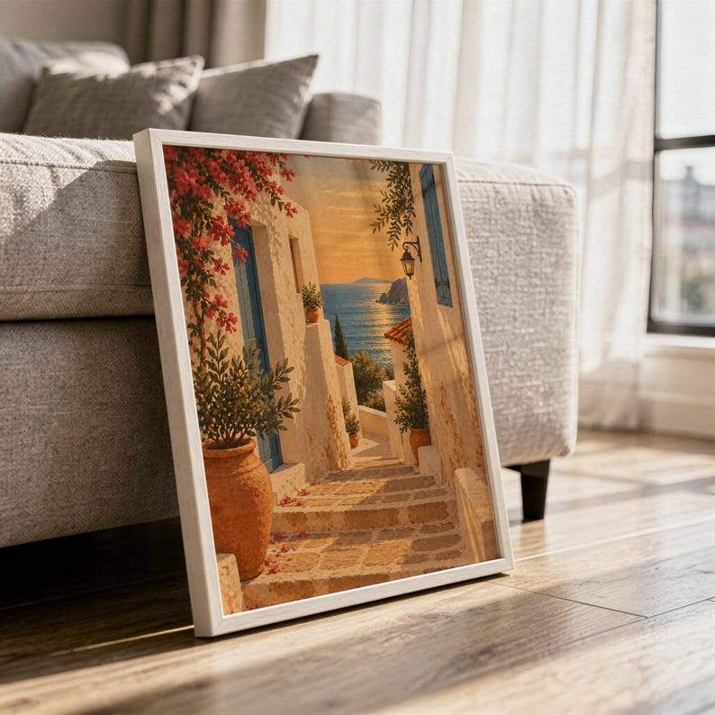 Mediterranean coastal village wall art print in a white frame leaning against a gray sofa on a sunlit wooden floor.