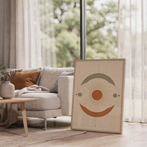 Abstract sun wall art print leaning on the floor beside a cream armchair next to large garden windows.