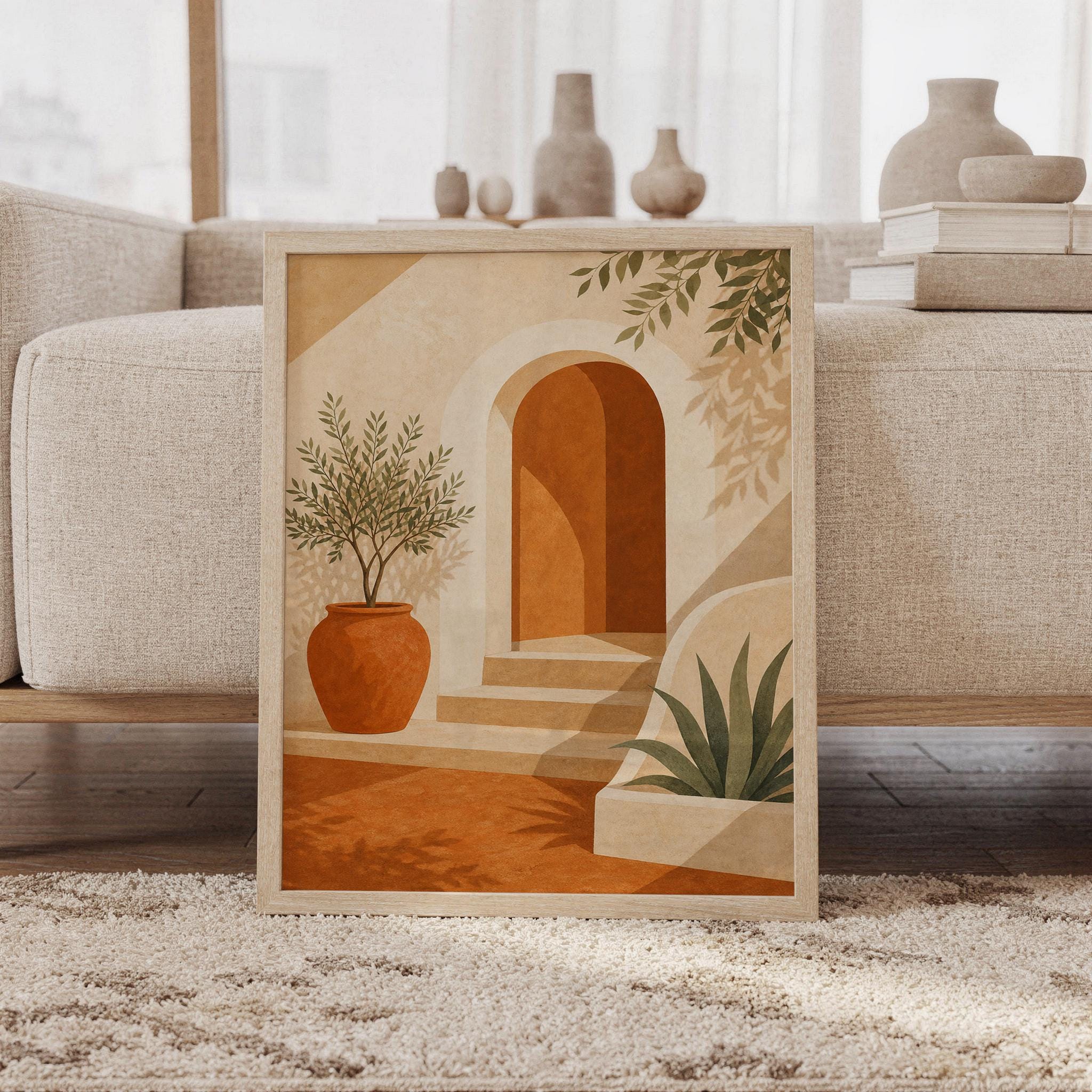 Mediterranean arch wall art print of a sunlit terracotta arch with a curved stairway, a clay pot and leafy plants in sand, cream and olive tones, framed and leaning against a beige sofa on a textured rug in a bright Scandinavian living room.
