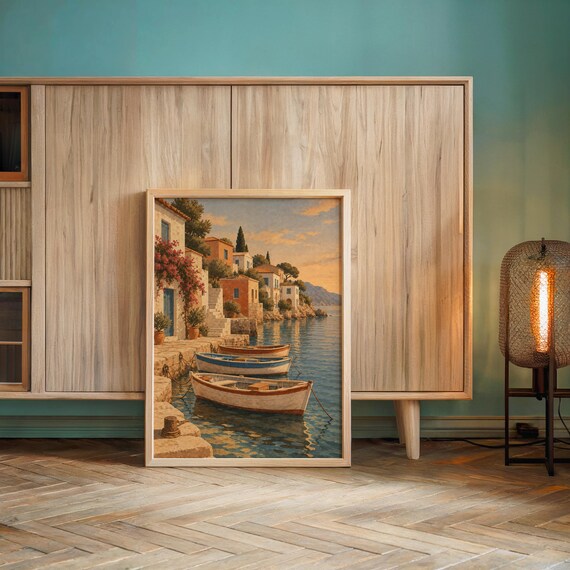 Mediterranean coastal village wall art print leaning against a wooden cabinet by a teal wall beside a glowing floor lamp.
