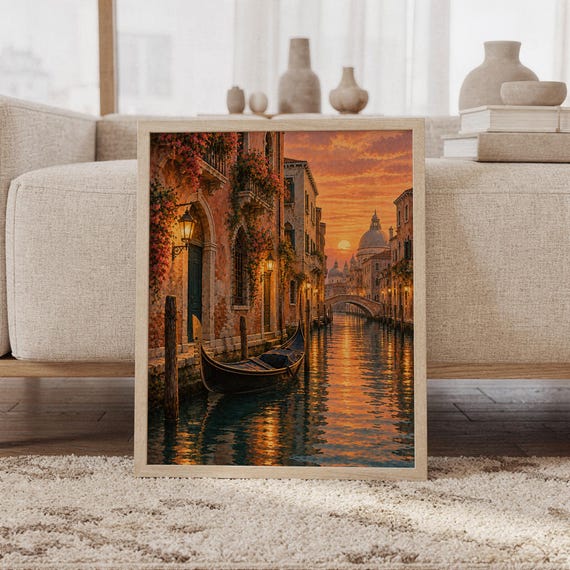 Venice canal sunset wall art print featuring glowing golden orange and peach skies, historic Venetian architecture, flower-covered facades, a gondola, and shimmering canal reflections in a painterly cityscape style, framed and leaning against a beige sofa on a textured rug in a bright Scandinavian living room.