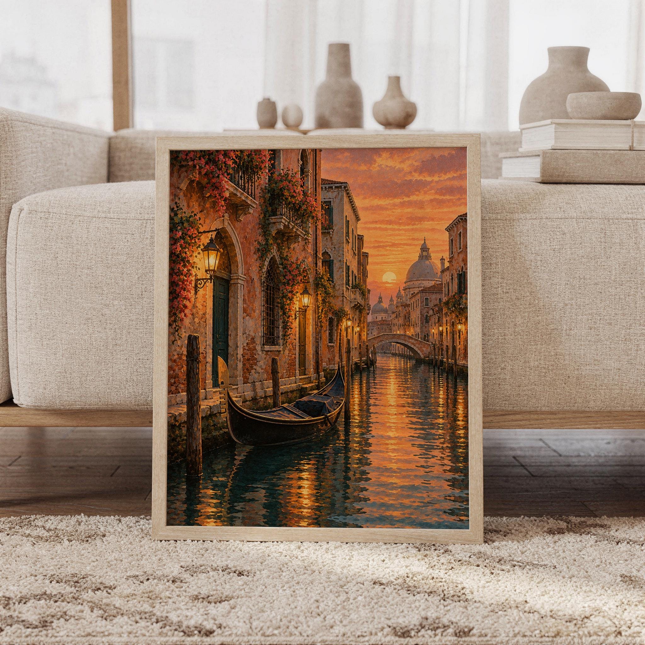 Venice canal sunset wall art print featuring glowing golden orange and peach skies, historic Venetian architecture, flower-covered facades, a gondola, and shimmering canal reflections in a painterly cityscape style, framed and leaning against a beige sofa on a textured rug in a bright Scandinavian living room.