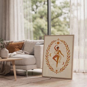 Framed neutral boho dancer art print leaning on the carpet next to a light wood coffee table and cozy living room sofa.