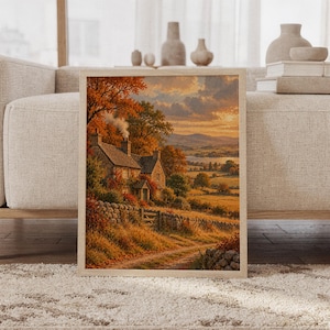 Autumn cottage landscape wall art print featuring glowing burnt orange foliage, warm golden fields, a stone cottage, rolling hills, and a painterly countryside style, framed and leaning against a beige sofa on a textured rug in a bright Scandinavian living room.