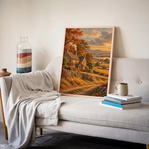 Autumn cottage landscape wall art print leaning on a cream sofa against a beige wall beside books and a mug.
