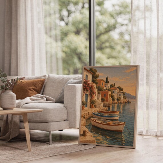 Mediterranean coastal village wall art print leaning on the floor beside a cream armchair next to large garden windows.