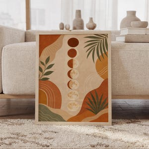 Boho moon phases wall art print of a vertical row of moon phases with abstract organic shapes and palm leaves in terracotta, sage green and beige, framed and leaning against a beige sofa on a textured rug in a bright Scandinavian living room.