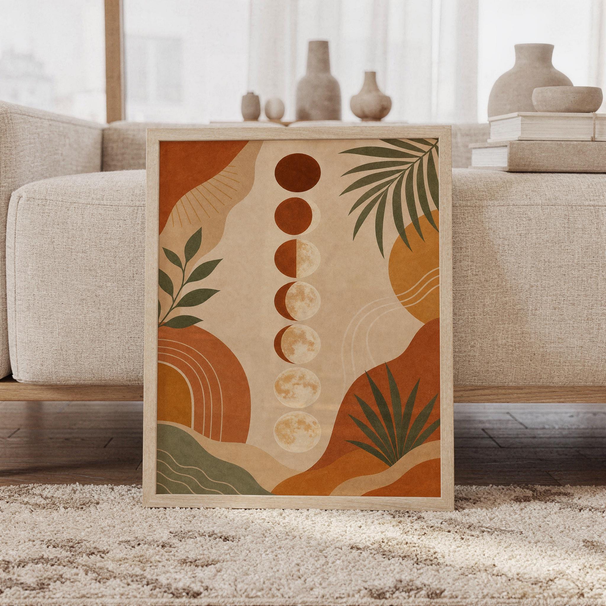 Boho moon phases wall art print of a vertical row of moon phases with abstract organic shapes and palm leaves in terracotta, sage green and beige, framed and leaning against a beige sofa on a textured rug in a bright Scandinavian living room.