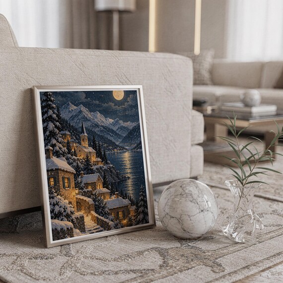 Snowy mountain village wall art print in a silver frame leaning against a sofa beside a decorative marble sphere.