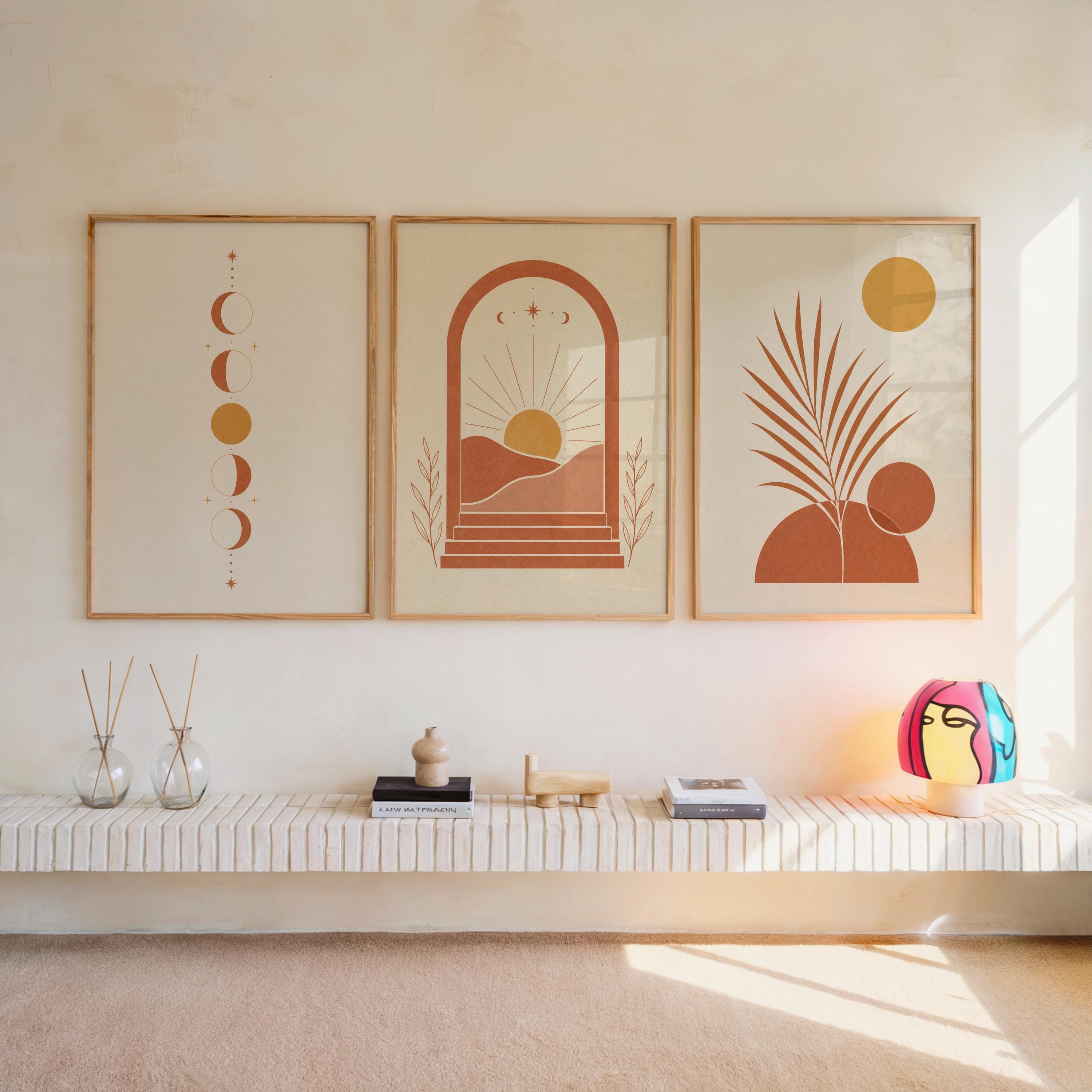 Set of 3 boho wall art prints featuring terracotta moon phases, abstract sun arch, and botanical palm leaf illustrations framed symmetrically on a warm sunlit wall above a floating wooden shelf with decorative items in a modern living room.