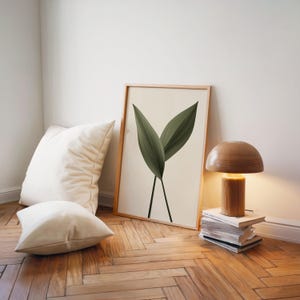 A framed print of two dark green leaves on a light background leans against a white wall, next to a wooden lamp and white pillows on a wooden floor.