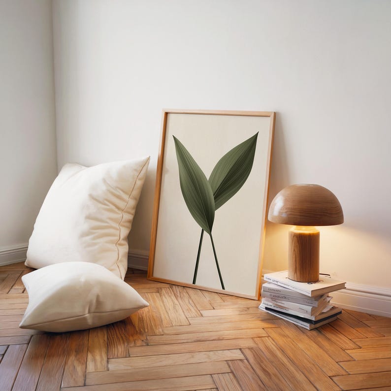 A framed print of two dark green leaves on a light background leans against a white wall, next to a wooden lamp and white pillows on a wooden floor.