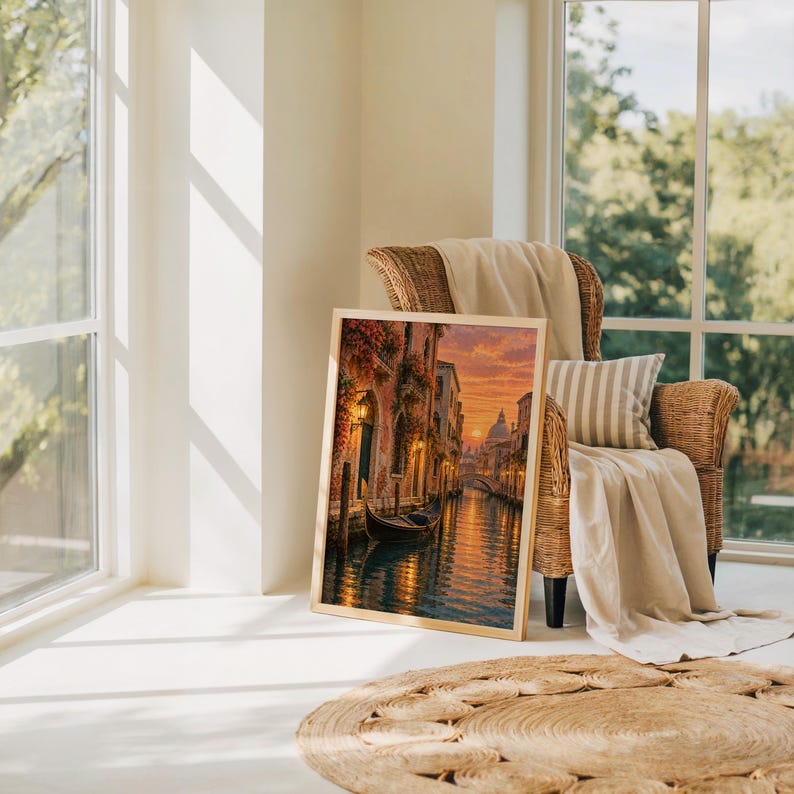 Venice canal sunset wall art print leaning on the floor beside a rattan armchair and jute rug in a bright sunlit room.