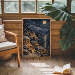 Snowy mountain village wall art print leaning against warm wood paneling beside a wooden armchair and a monstera plant.