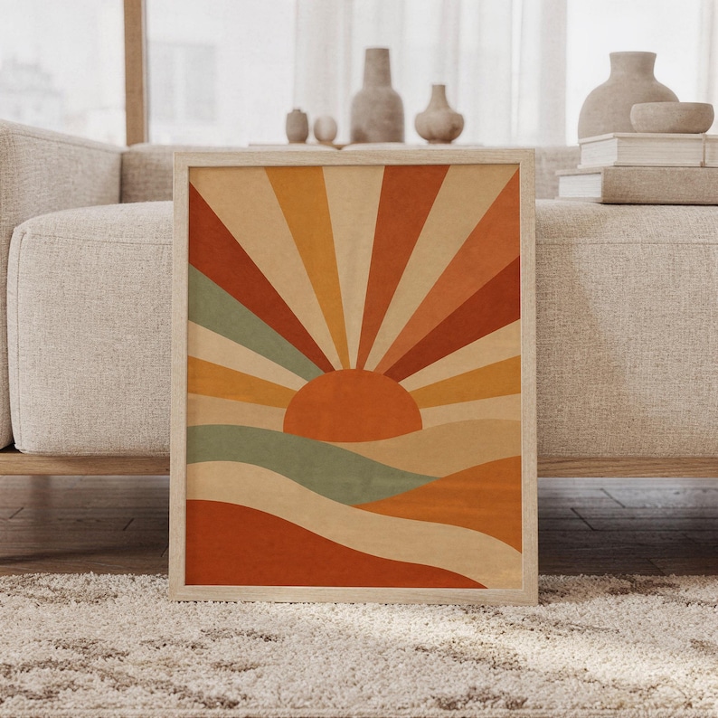 Retro sunrise wall art print of a mid-century geometric sun with bold rays rising over rolling hills in terracotta, burnt orange, ochre and sage, framed and leaning against a beige sofa on a textured rug in a bright Scandinavian living room.