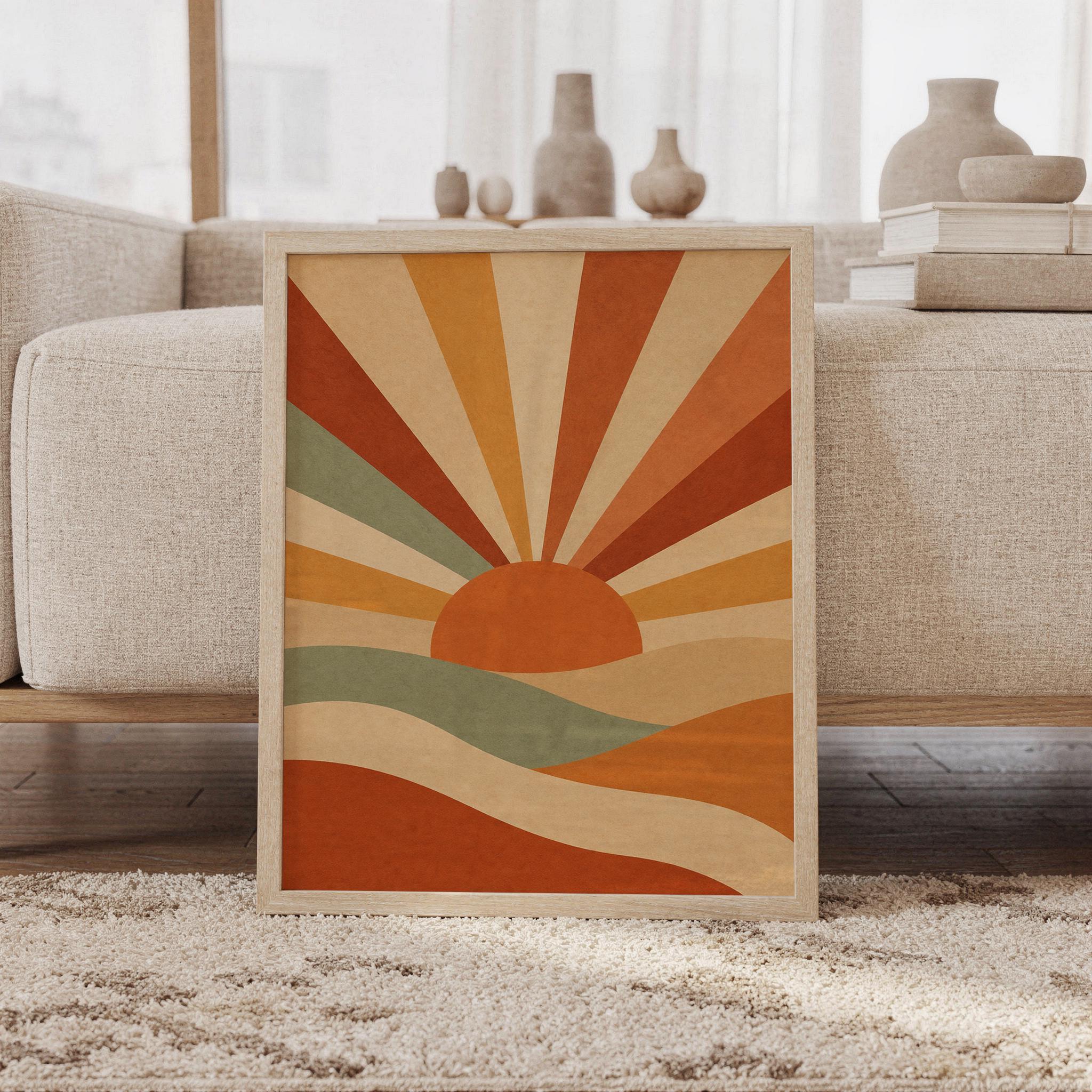 Retro sunrise wall art print of a mid-century geometric sun with bold rays rising over rolling hills in terracotta, burnt orange, ochre and sage, framed and leaning against a beige sofa on a textured rug in a bright Scandinavian living room.