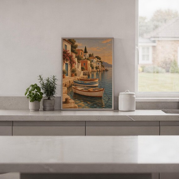 Mediterranean coastal village wall art print standing on a white kitchen counter beside potted herbs.