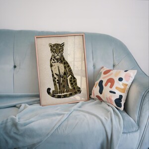 A framed print of a sitting leopard with dark green and brown spots on a light background, leaning against a light blue velvet couch with a patterned throw pillow.