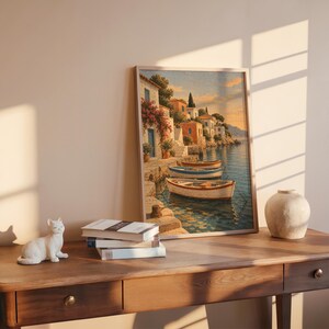 Mediterranean coastal village wall art print leaning on a wooden desk against a warm beige wall in soft window sunlight.