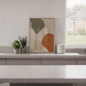 Framed abstract leaf art print resting on a white countertop with indoor plants.