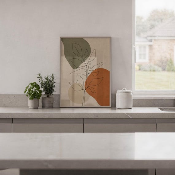 Framed abstract leaf art print resting on a white countertop with indoor plants.