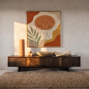 Abstract line art face print hanging above a dark wood console and jute rug in a warm neutral interior.