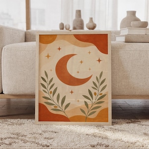 Celestial moon wall art print of a terracotta crescent moon with golden stars and olive branches over abstract cream, ochre and rust shapes, framed and leaning against a beige sofa on a textured rug in a bright Scandinavian living room.