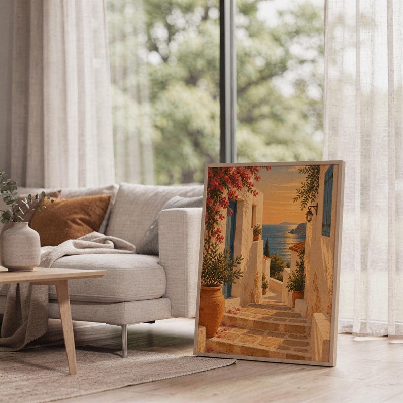 Mediterranean coastal village wall art print leaning on the floor beside a cream armchair next to large garden windows.