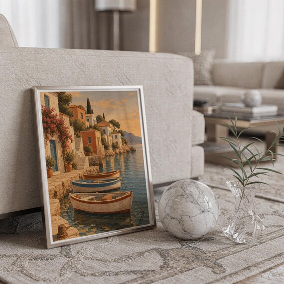 Mediterranean coastal village wall art print in a silver frame leaning against a sofa beside a decorative marble sphere.